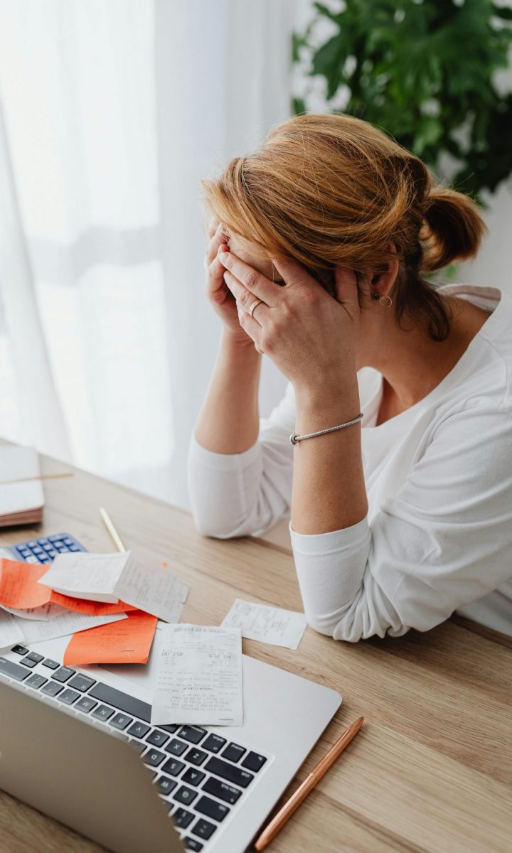 Overwhelmed woman sitting at desk with laptop and documents, showing stress and frustration.